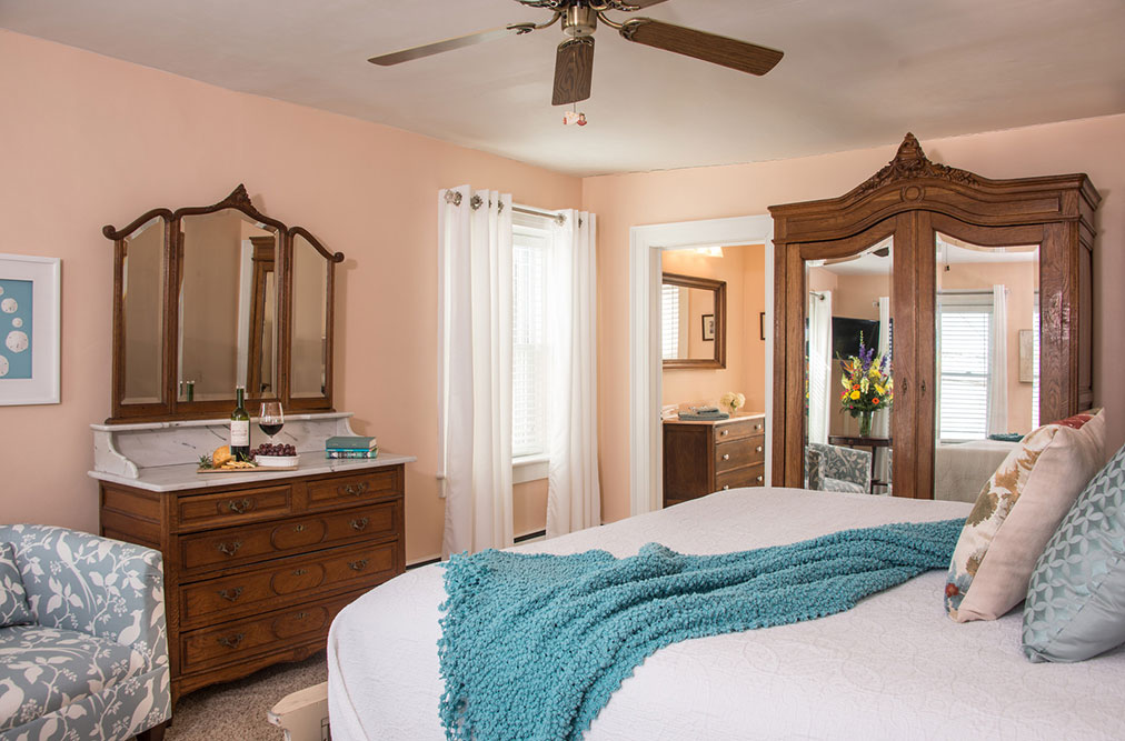 A light peach bedroom featuring a king bed with white linens and a teal throw blanket. The room includes a large antique oak armoire with mirrored doors, a matching wooden dresser with a tri-fold mirror, and a glimpse of an ensuite bathroom through an open doorway.