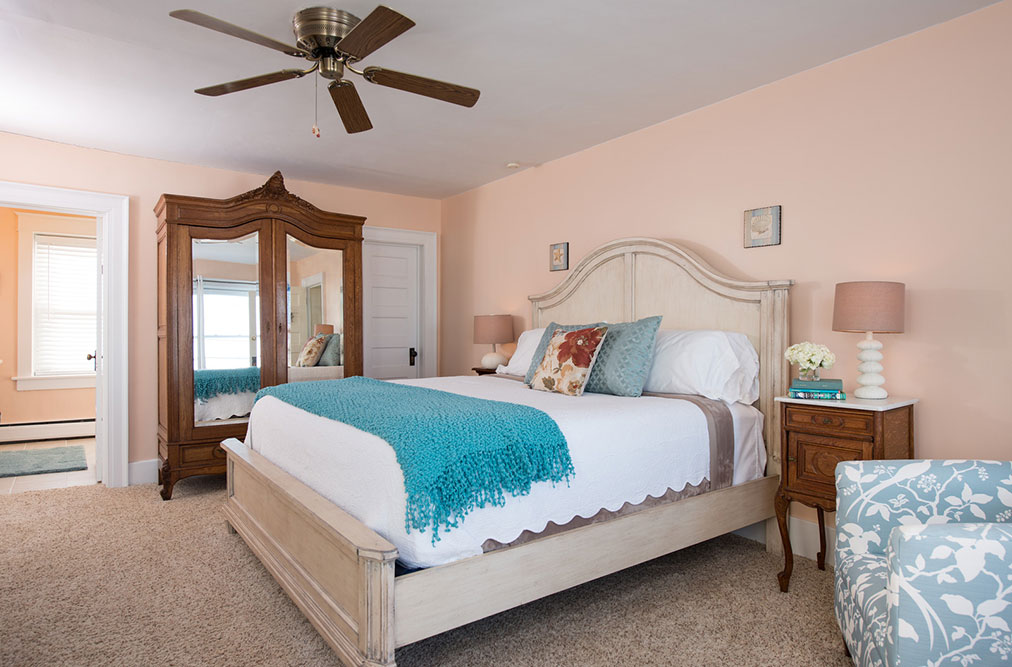 A brightly lit bedroom with soft peach walls featuring a white king-size bed with a teal throw blanket. To the left, a large antique wooden armoire with mirrored doors stands near an open doorway leading to a bathroom. A patterned light blue armchair is partially visible in the bottom right corner.