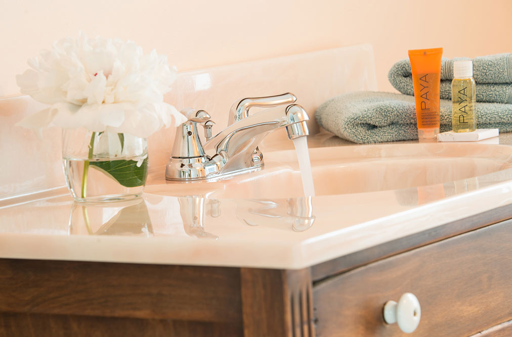 A close-up of a bathroom vanity with a running chrome faucet. On the counter, there are folded teal towels, orange "PAYA" brand toiletries, and a small glass vase containing a white peony.