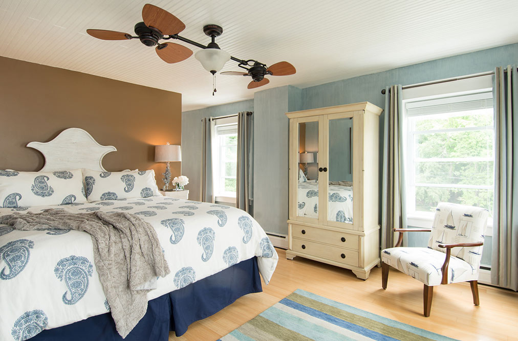 A light-filled bedroom with soft blue walls and a unique multi-blade ceiling fan. It features a king-size bed with a blue and white paisley comforter, a large cream-colored mirrored armoire, and a matching patterned accent chair.