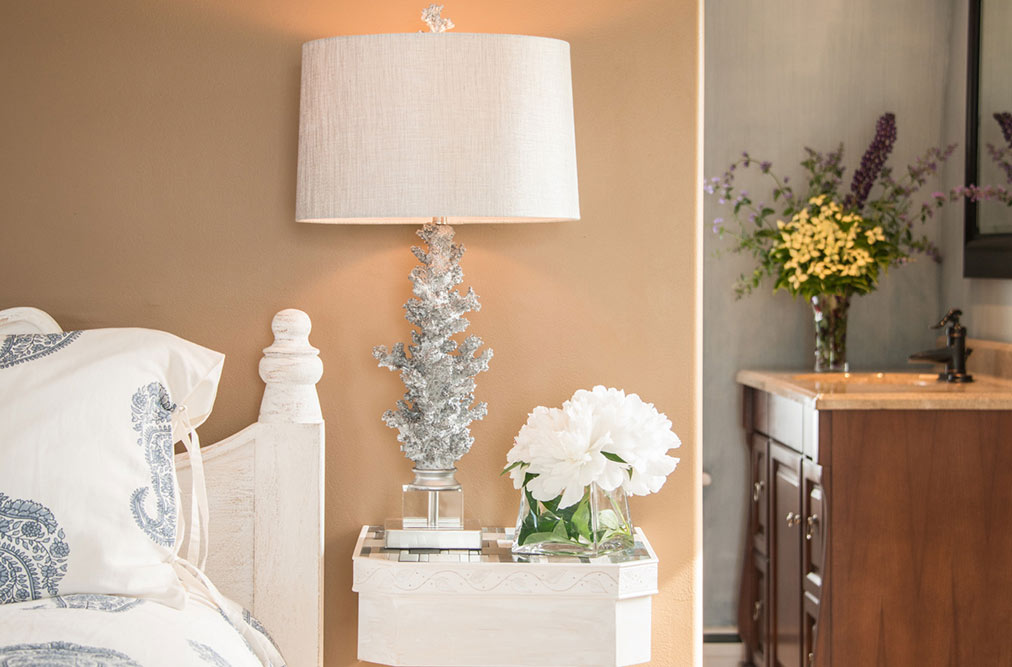 A close-up of a bedside table featuring a silver coral-inspired lamp with a white shade. Next to the lamp is a small glass vase with white hydrangeas, while a large bouquet of yellow and purple wildflowers is visible on a vanity in the background.