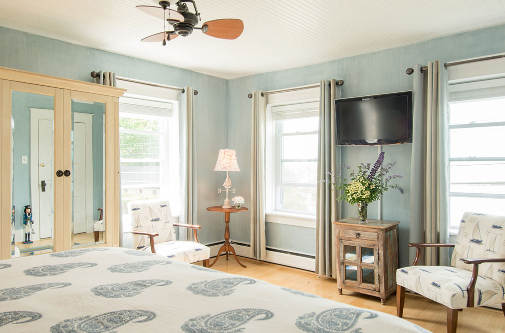 A light blue bedroom featuring two patterned armchairs and a small round wooden table with a lamp. A flat-screen TV is mounted on the wall above a wooden cabinet topped with a large flower arrangement, and three windows with white blinds let in natural light.