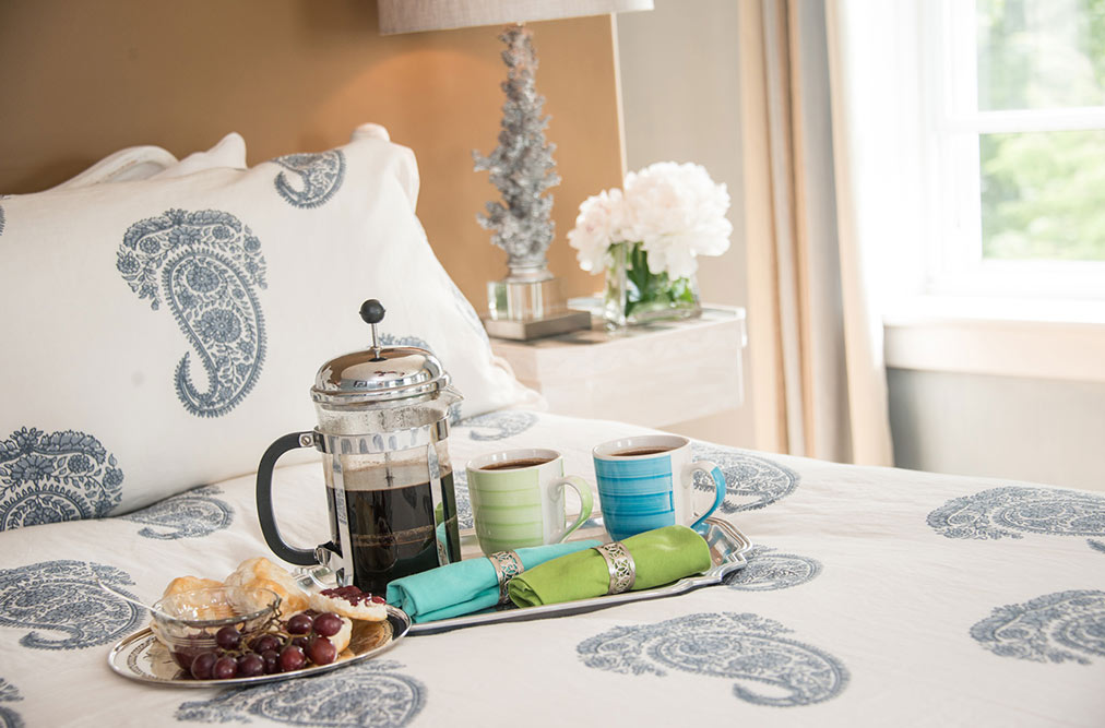 A close-up of a silver tray resting on a blue and white paisley bedspread. The tray holds a French press coffee maker, two colorful mugs, teal and lime green napkins, and a small plate of pastries and grapes. In the background, a white bedside table holds a coral-inspired lamp and white hydrangeas.