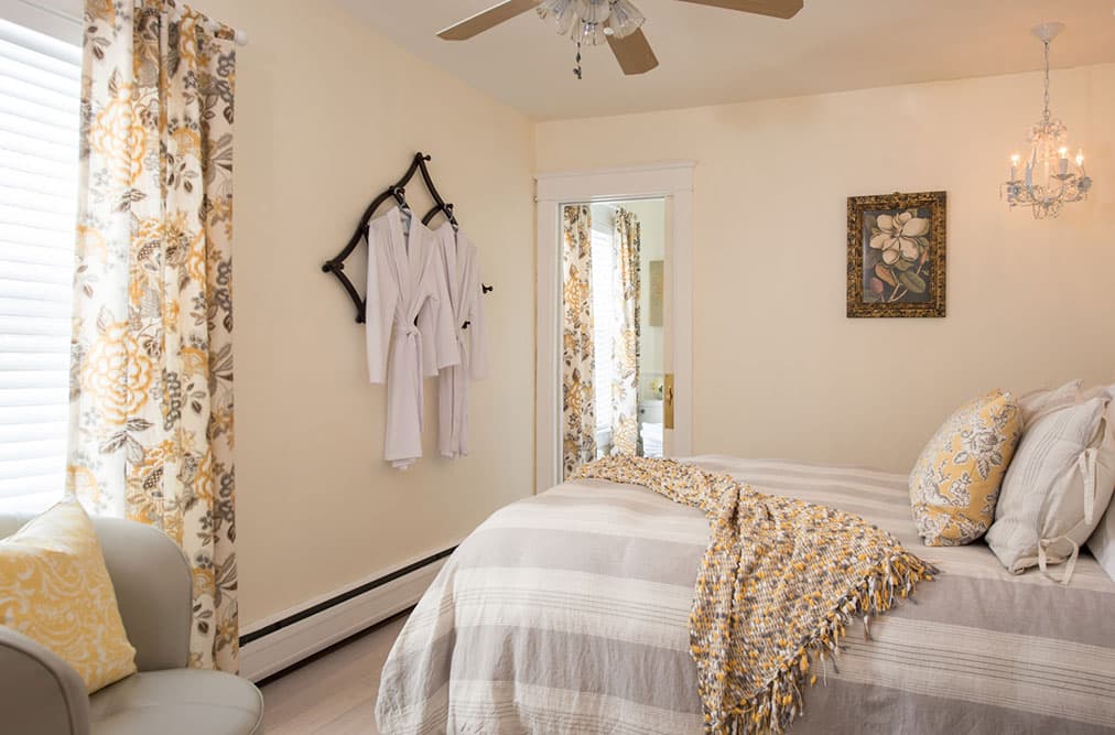 A cozy bedroom with creamy walls and yellow accents, featuring a queen bed with a grey and white striped comforter, two white robes hanging on a wall-mounted rack, and a small crystal chandelier.