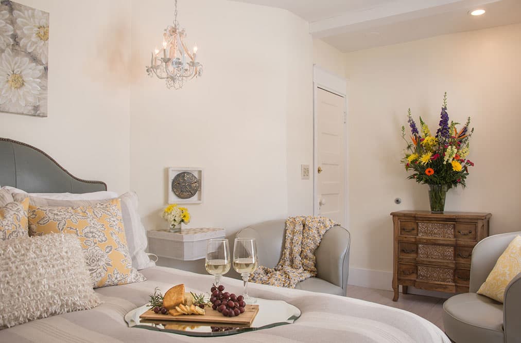 A bedroom sitting area featuring a grey armchair with a yellow pillow and a matching grey loveseat. In the foreground, a wooden tray on the bed holds white wine, cheese, and grapes, while a wooden dresser in the corner displays a large, colorful floral arrangement.