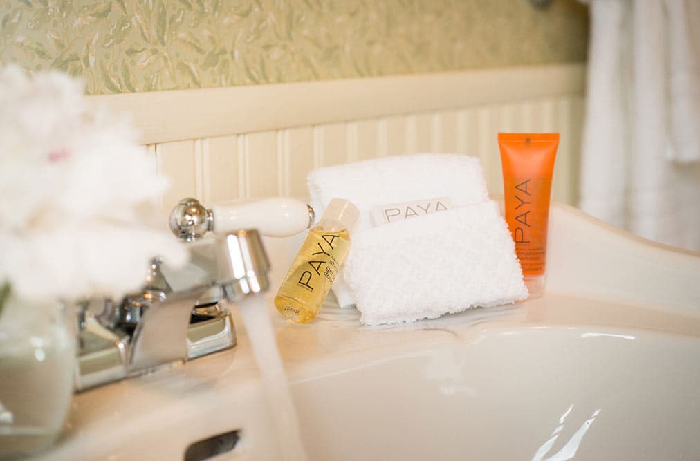 A close-up of a bathroom sink featuring a chrome faucet with water running. Resting on the ledge are two small tubes of orange "PAYA" brand toiletries and a folded white washcloth embroidered with "LAKESIDE."