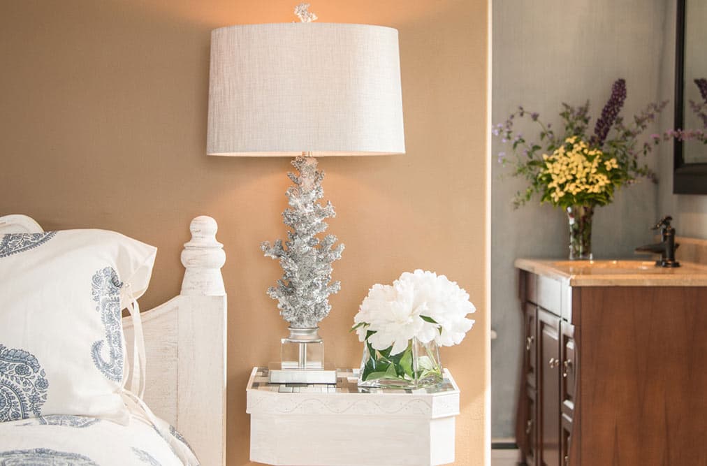 A close-up of a bedside table featuring a silver coral-inspired lamp with a white shade. Next to the lamp is a small glass vase with white hydrangeas, while a large bouquet of yellow and purple wildflowers is visible on a vanity in the background.