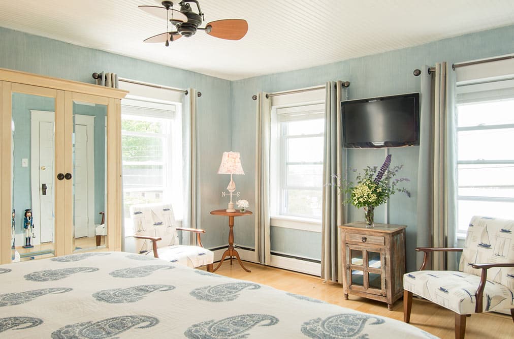 A light blue bedroom featuring two patterned armchairs and a small round wooden table with a lamp. A flat-screen TV is mounted on the wall above a wooden cabinet topped with a large flower arrangement, and three windows with white blinds let in natural light.