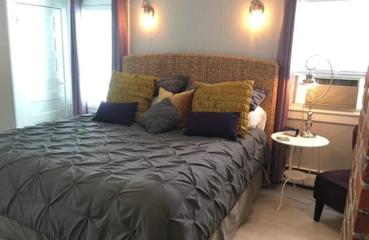 A king-size bed with a dark gray quilted comforter, featuring yellow and dark purple accent pillows. The room has light gray walls, a wicker headboard, and two bedside lamps casting a warm glow.