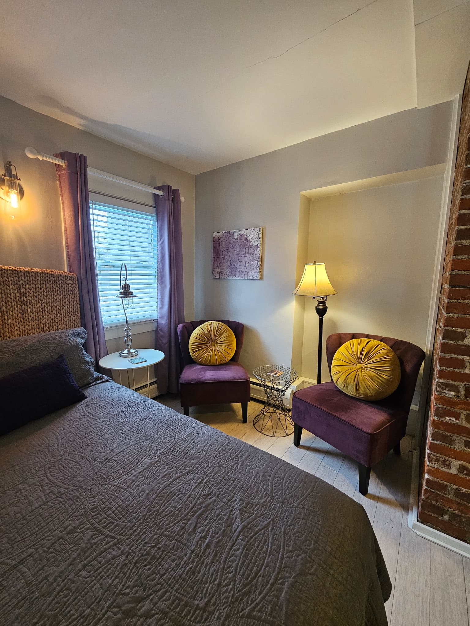 Two purple accent chairs with yellow circular pillows positioned in a corner next to a bed with a gray quilted comforter. The room features light gray walls, a floor lamp, a small round side table, and a partial view of an exposed brick wall.