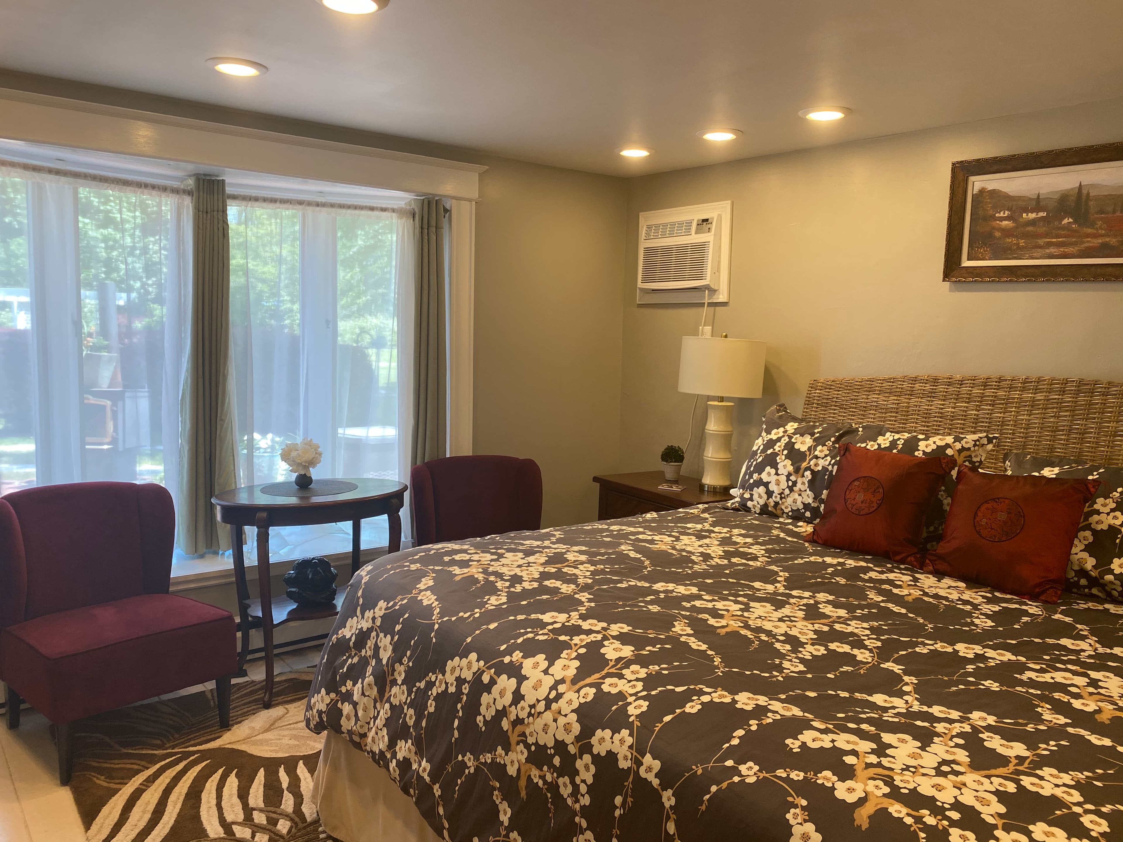 A bedroom with grey walls and a large bow window, featuring a king bed with a floral cherry blossom bedspread, two red accent pillows, and two dark purple velvet chairs near a small wooden table.