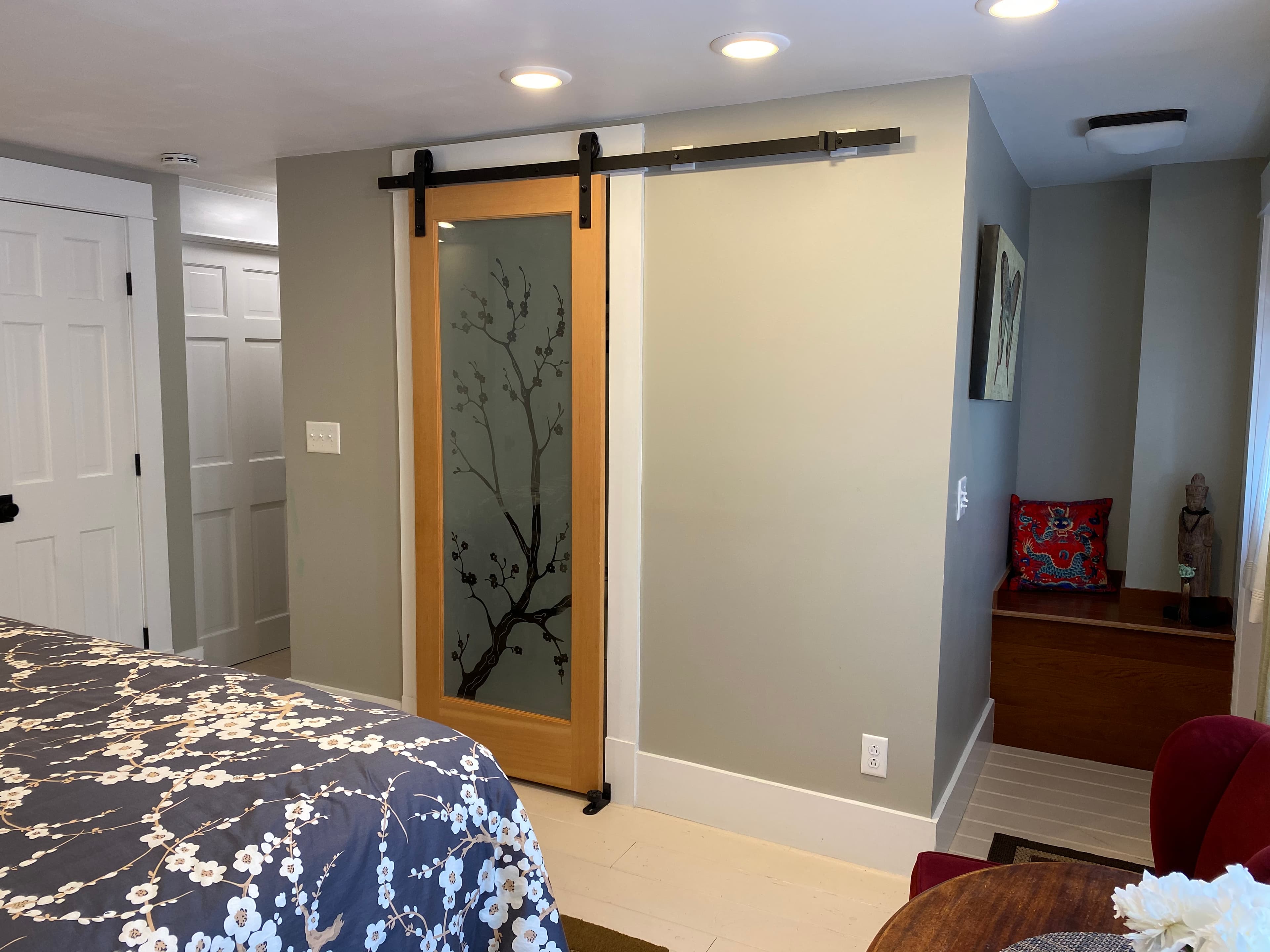 A sliding wooden barn door with a frosted glass pane featuring a black cherry blossom branch design, set against light grey walls next to a bed with a floral comforter and a small wooden storage bench.