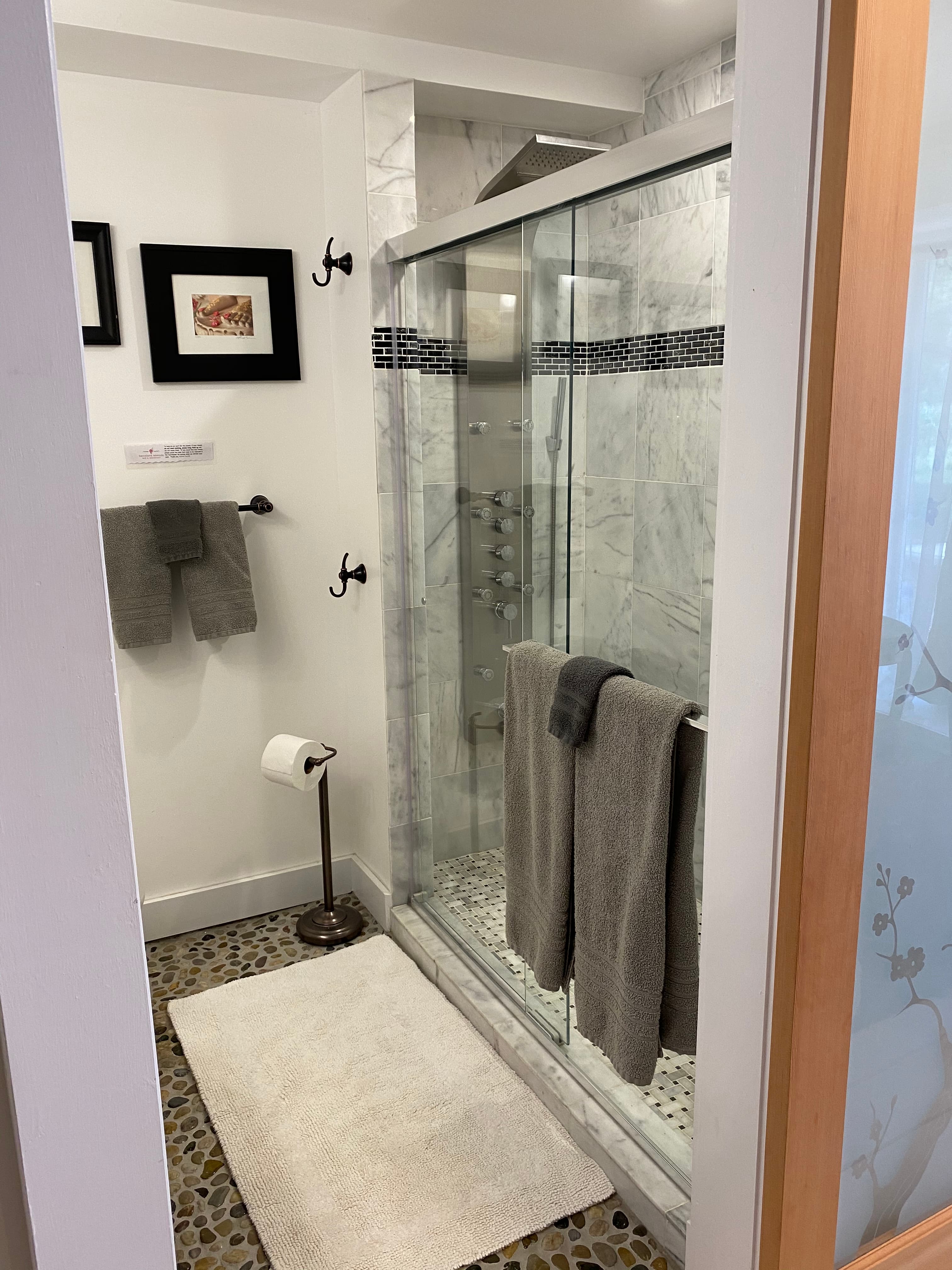 A modern spa bathroom featuring a large walk-in shower with glass doors, white marble-style wall tiles, a pebble-textured floor, and plush grey towels hanging on a silver handle.