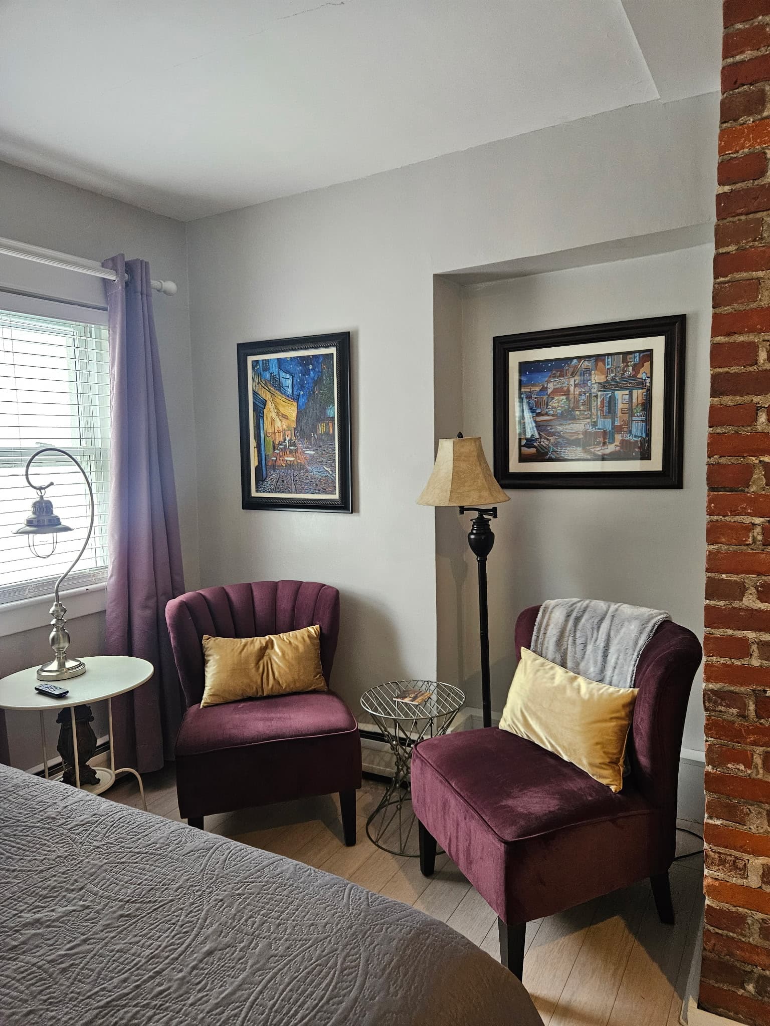Two purple accent chairs with yellow circular pillows positioned in a corner next to a bed with a gray quilted comforter. The room features light gray walls, a floor lamp, a small round side table, and a partial view of an exposed brick wall.
