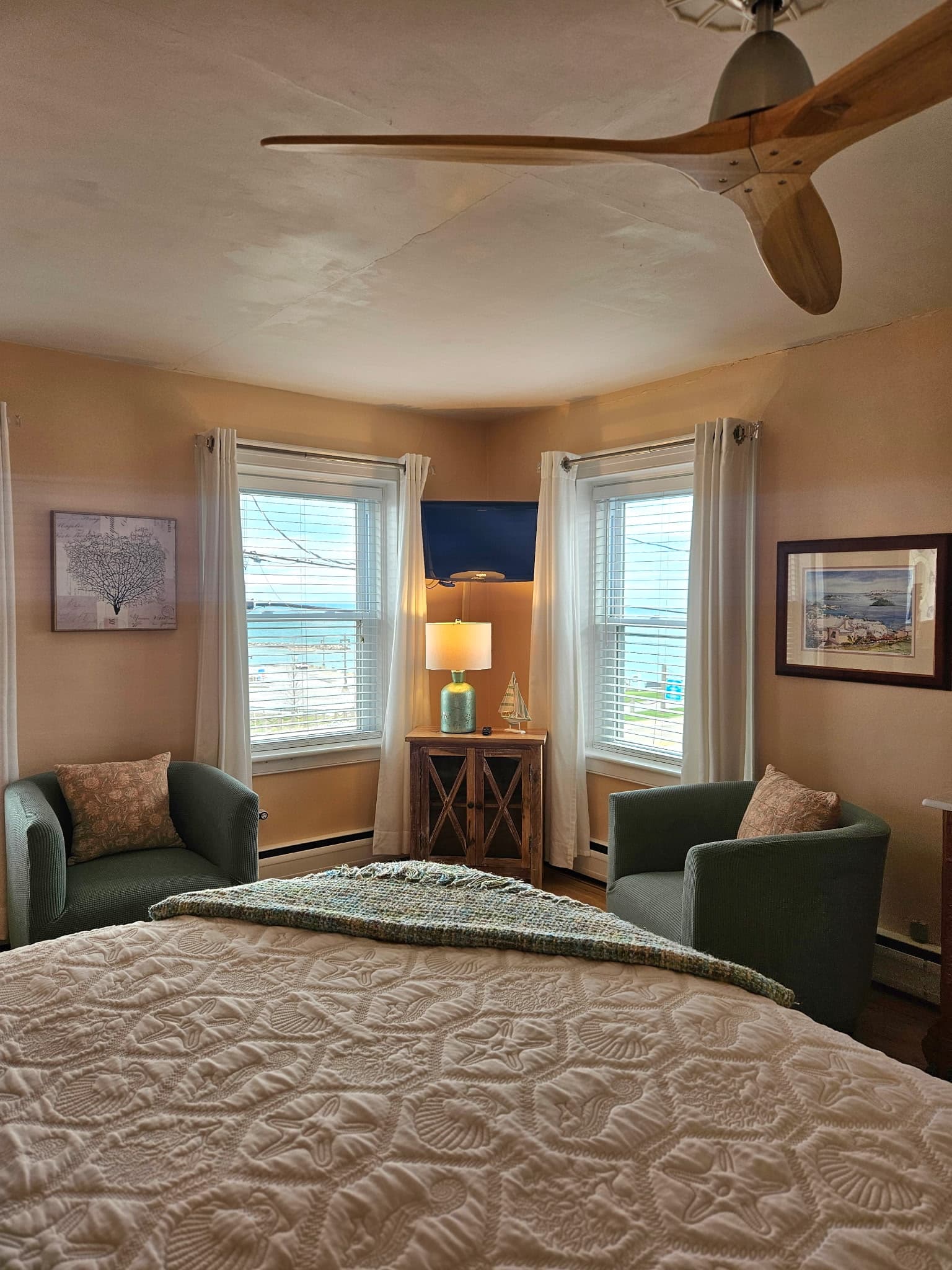 A cozy sitting area in a peach-colored room featuring two blue floral armchairs angled toward a small round wooden table with a large, colorful flower arrangement. White sheer curtains frame the windows, and a flat-screen TV is mounted in the corner.