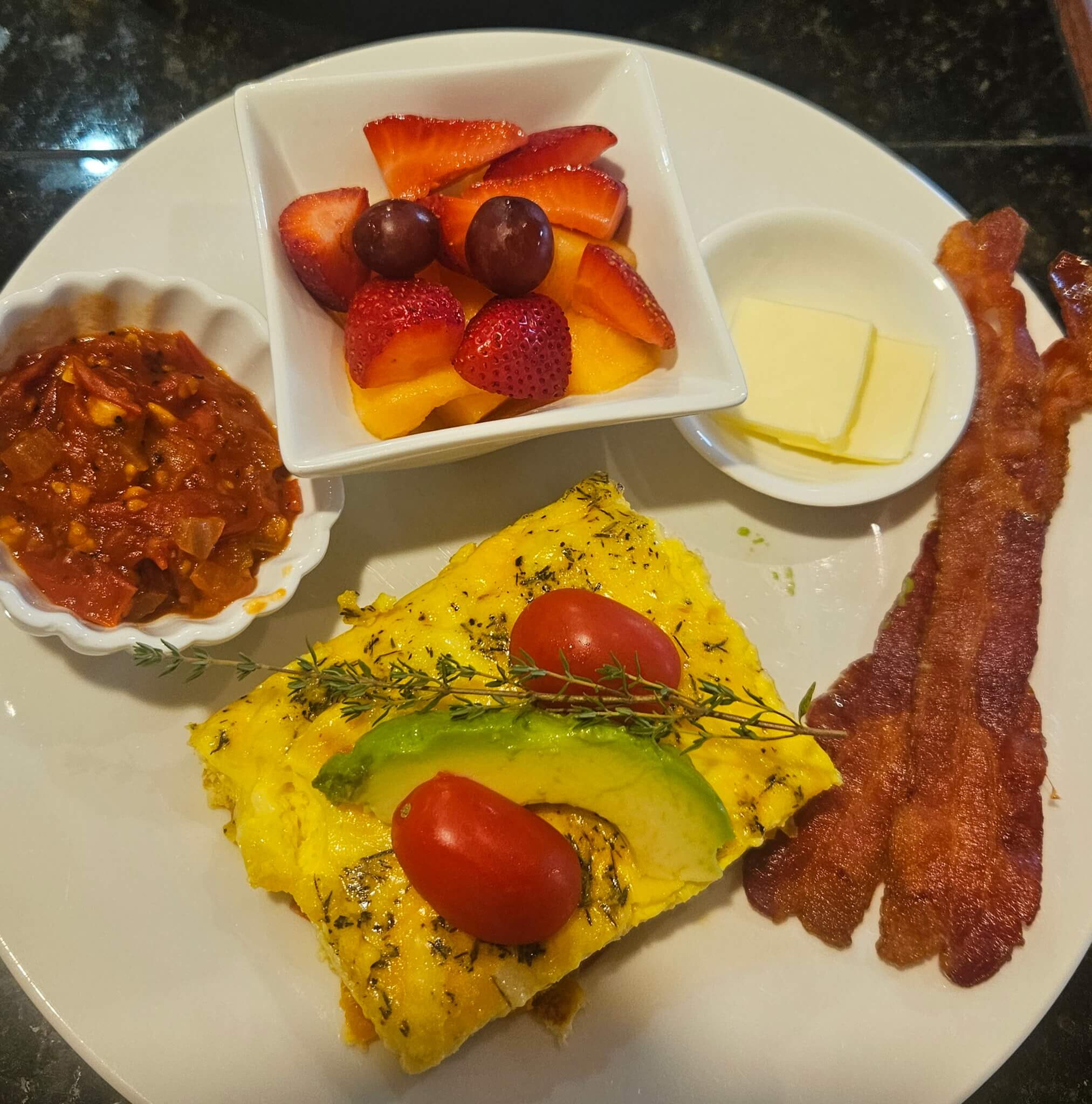 A plate featuring an omelette topped with tomatoes and avocado, a side of bacon, butter, a fruit salad, and a serving of salsa.