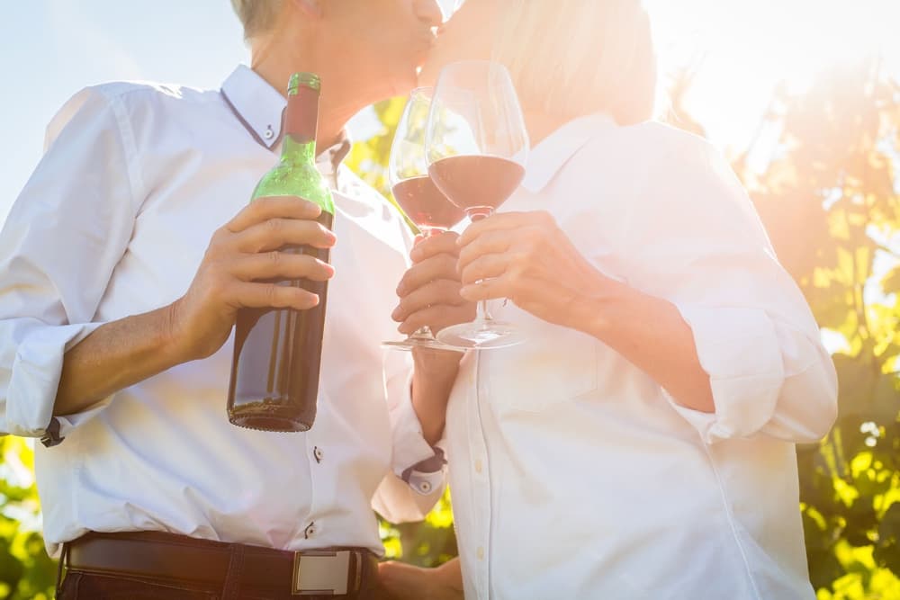 A couple shares a kiss while holding wine glasses and a bottle in a sunlit vineyard.
