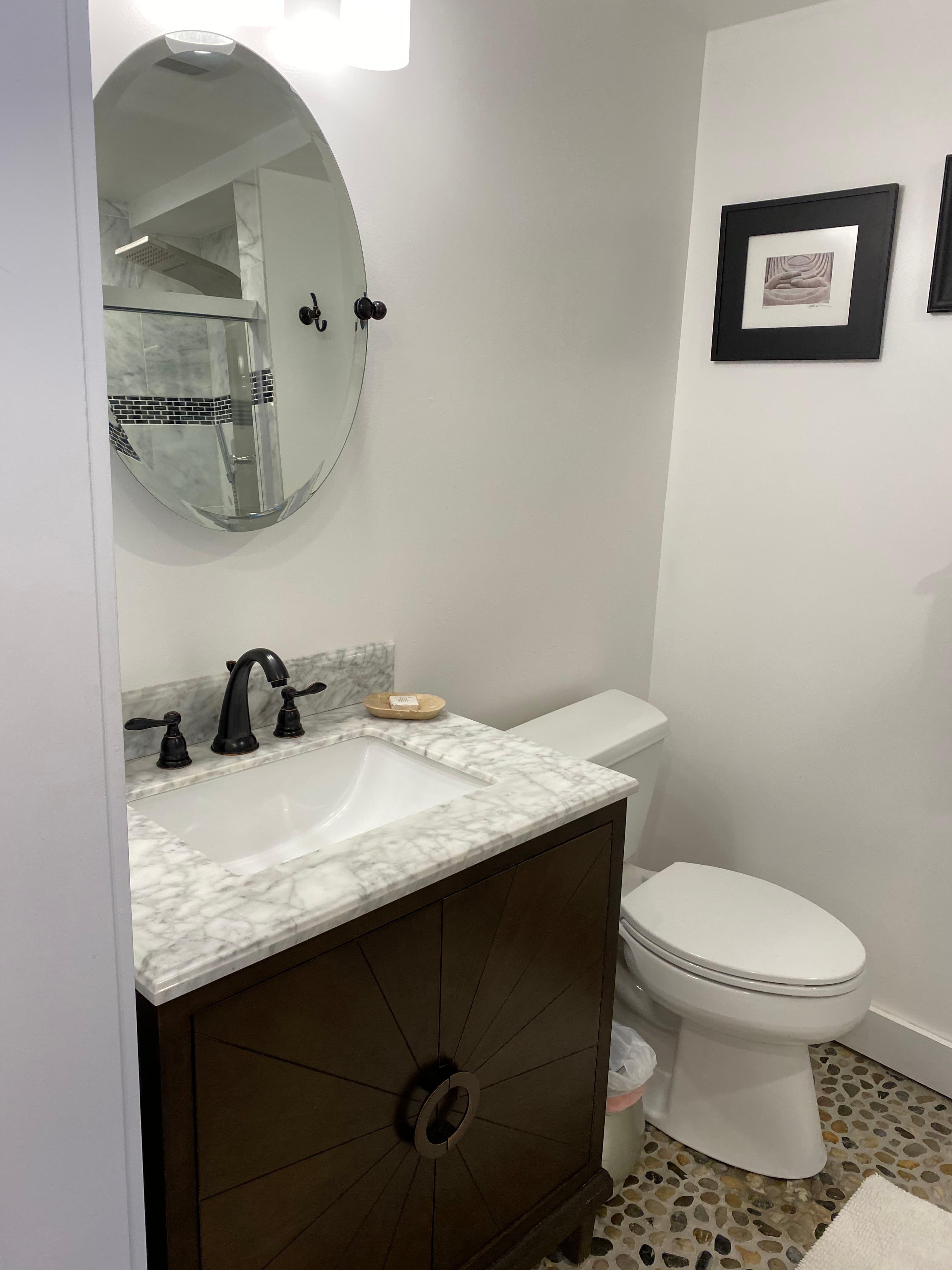 A bathroom vanity with a white marble top and dark wood cabinet featuring a sunburst design, next to a white toilet. An oval mirror hangs above the sink, reflecting a walk-in shower.