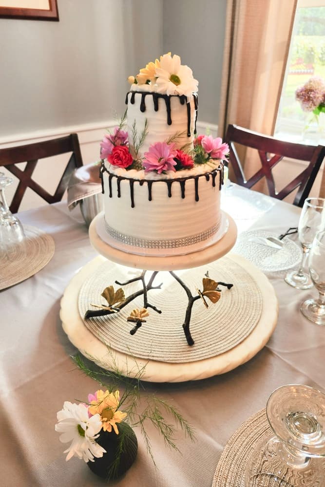 A beautifully decorated two-tier cake adorned with flowers and chocolate drizzle on a stylish cake stand.