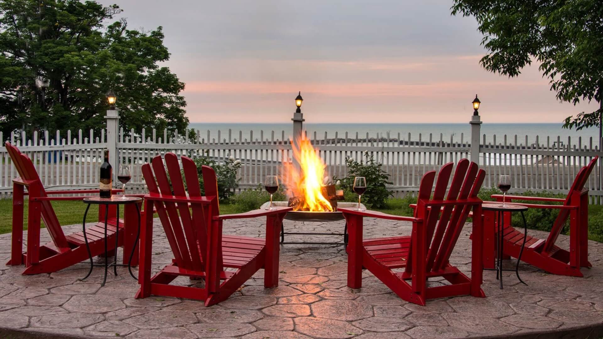 A cozy patio scene with red Adirondack chairs surrounding a fire pit, overlooking a serene lake at sunset.