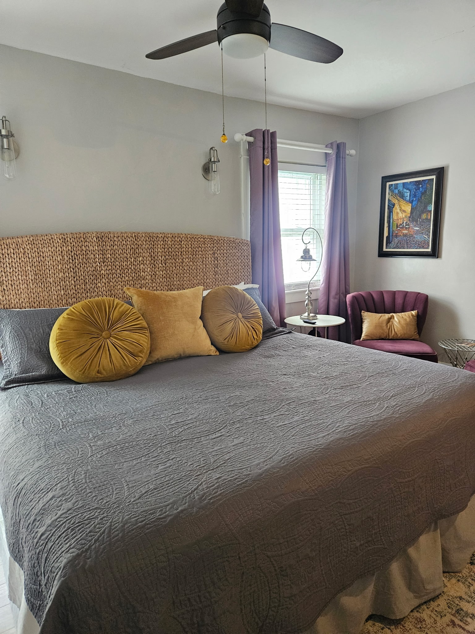A king-size bed with a dark gray quilted comforter, featuring yellow and dark purple accent pillows. The room has light gray walls, a wicker headboard, and two bedside lamps casting a warm glow.