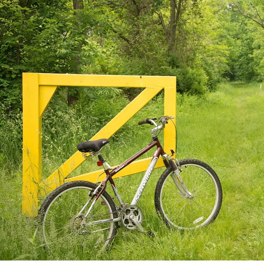 A maroon bike leans against a bright yellow gate on a grassy path.
