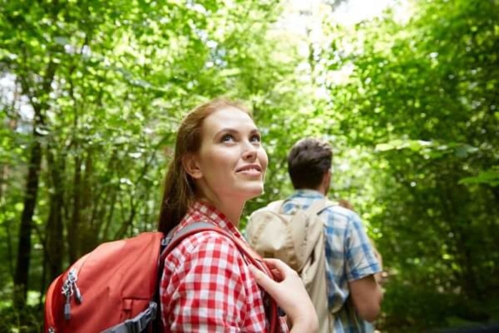A woman with a backpack smiles while walking in a lush green forest with a companion.