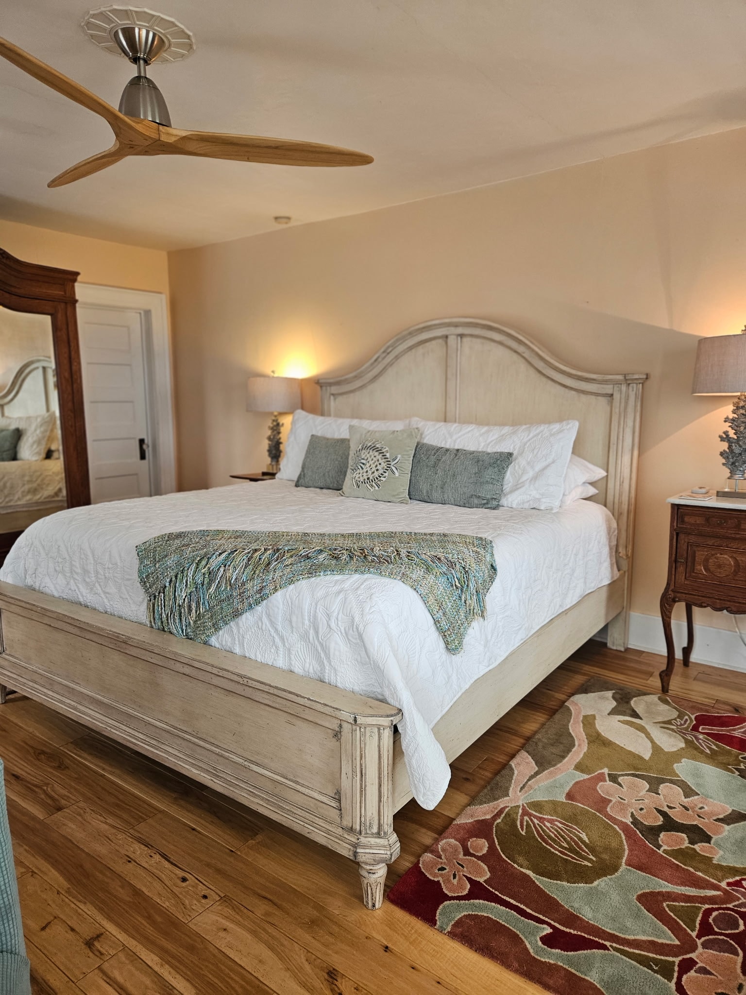 A light peach bedroom featuring a king bed with white linens and a teal throw blanket. The room includes a large antique oak armoire with mirrored doors, a matching wooden dresser with a tri-fold mirror, and a glimpse of an ensuite bathroom through an open doorway.