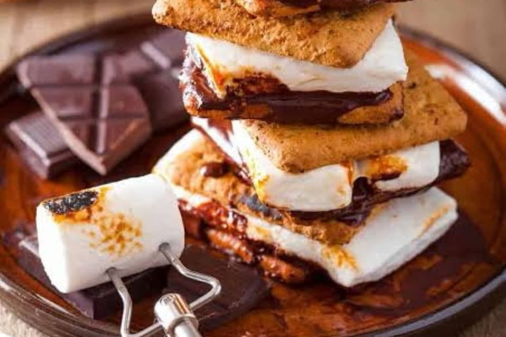 A stack of s'mores with toasted marshmallows, chocolate, and graham crackers, beside a roasting fork and pieces of chocolate on a wooden plate.