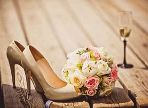 A pair of beige high-heeled shoes beside a wedding bouquet and a glass of champagne on a wooden surface.