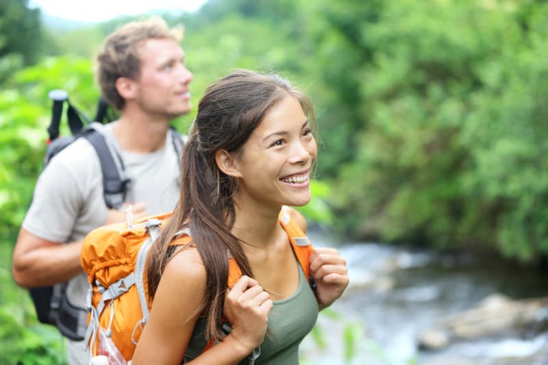 A smiling woman with a backpack hikes alongside a man in a lush, green outdoor setting.