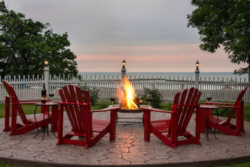 A cozy outdoor fire pit surrounded by red Adirondack chairs and a view of the water at dusk.