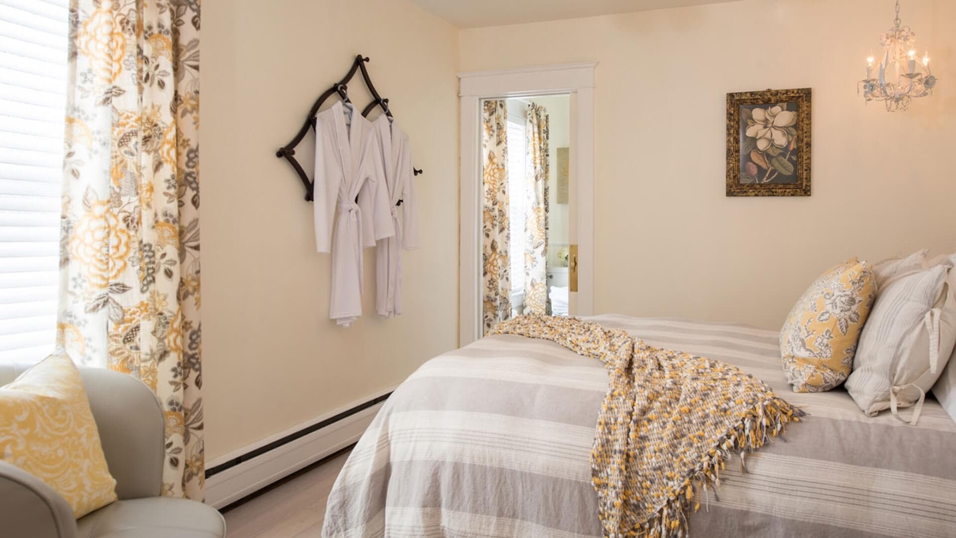 A cozy bedroom featuring a striped bedspread, patterned curtains, and white robes hanging on a wall.
