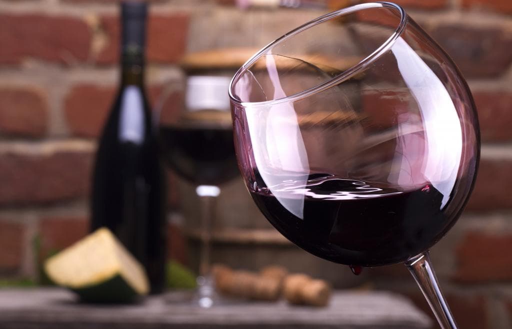 A close-up of a glass of red wine with blurred bottles and cheese in the background. A close-up of a glass of red wine with blurred bottles and cheese in the background.
