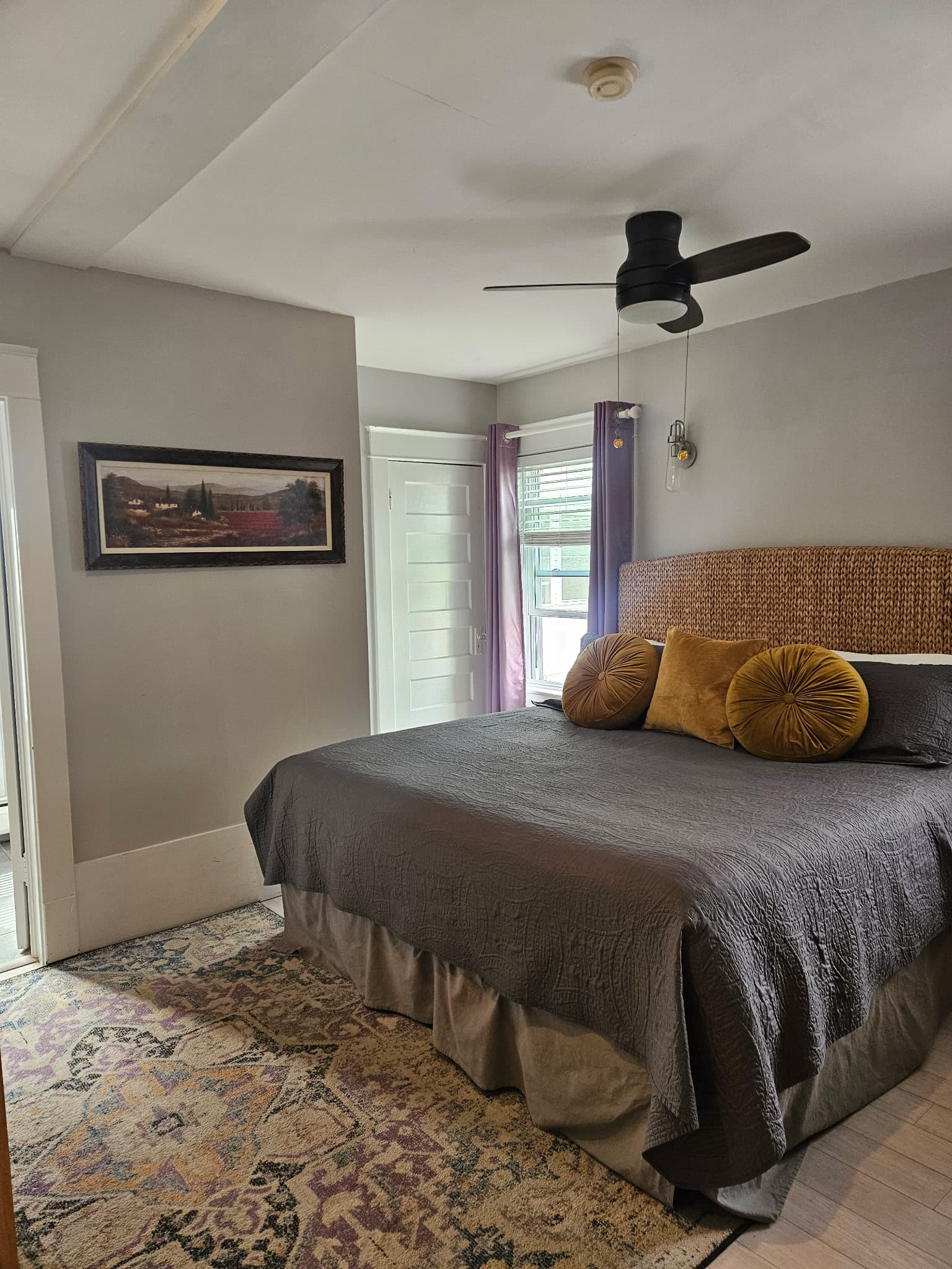 A cozy bedroom featuring a large bed with decorative pillows, a ceiling fan, and a window with light curtains.