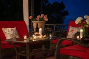 A cozy outdoor patio scene at night, featuring red chairs, a table with candles, wine, and flowers.