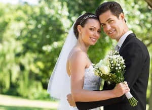A happy bride and groom pose together outdoors, smiling and holding a bouquet of flowers.