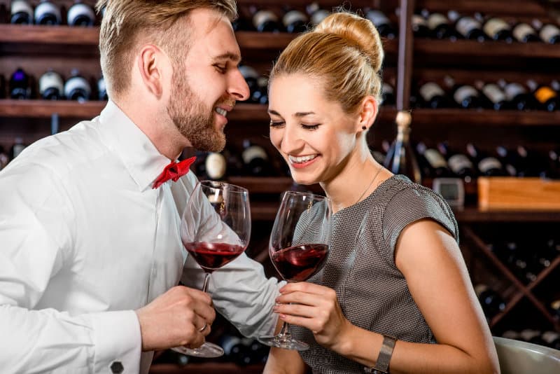 A man and woman smile at each other while clinking glasses of red wine in a wine cellar.
