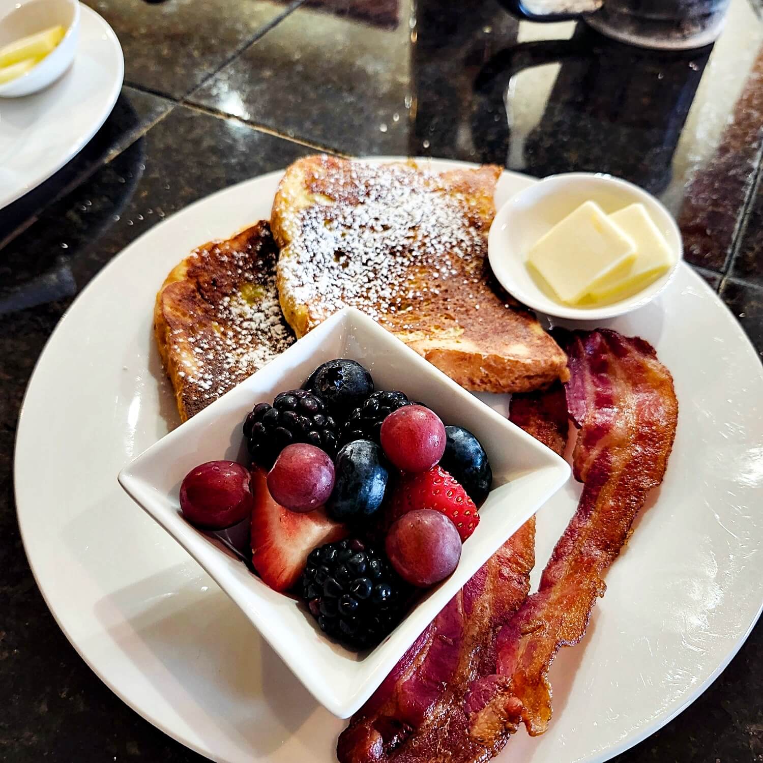 A plate of French toast dusted with powdered sugar, crispy bacon, and a bowl of mixed berries with a side of butter.