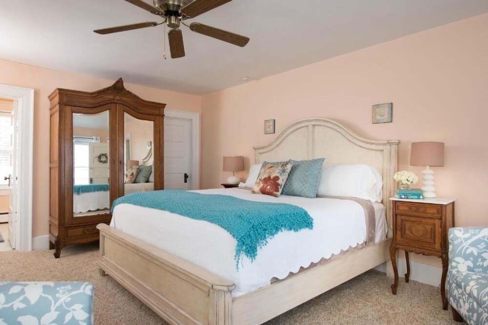 A cozy bedroom featuring a light-colored bed with a turquoise throw, vintage mirror, and warm peach walls.