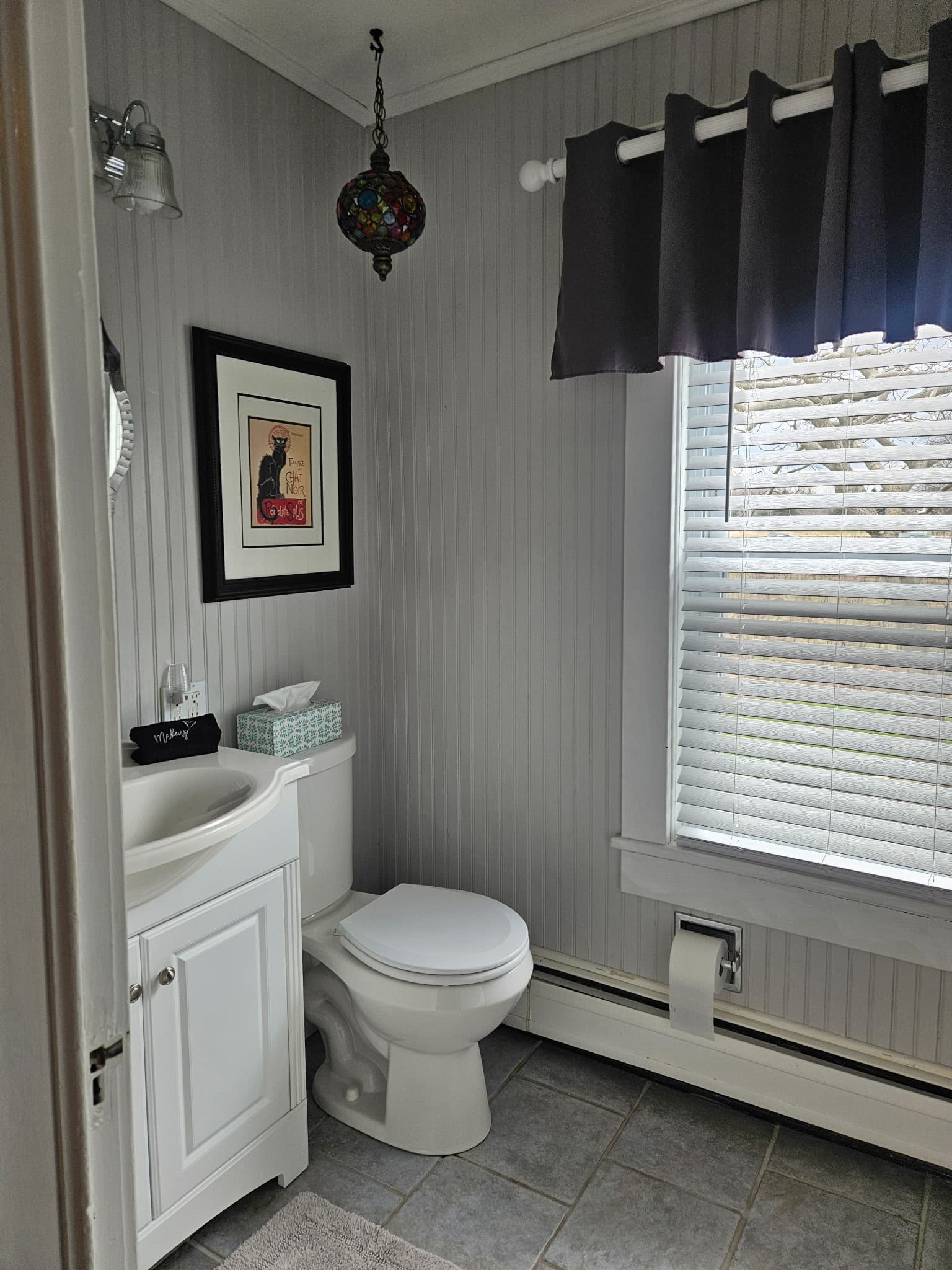 A modern bathroom featuring a toilet, sink, decorative artwork, and a window with blinds.